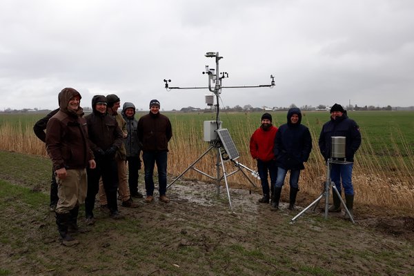 Eine Gruppe von Menschen steht neben einer Wetterstation auf dem Feld.