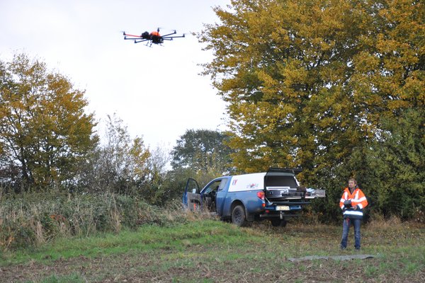 Eine Drohne fliegt an einem Knickrand. Rechts unten im Bild steht der Drohnenpilot mit Fernbedienung.