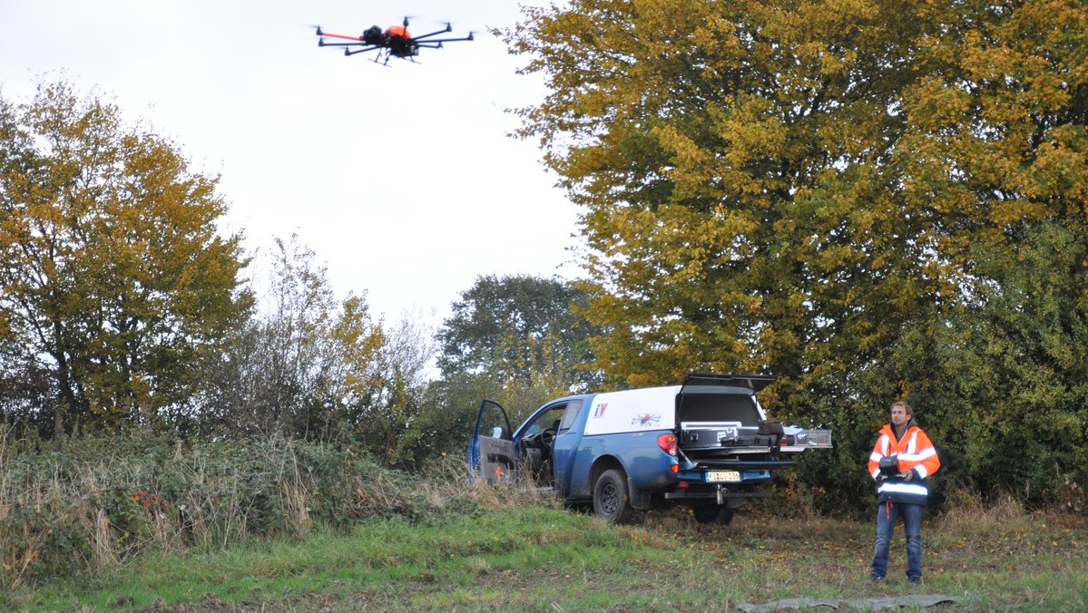 Eine Drohne fliegt an einem Knickrand. Rechts unten im Bild steht der Drohnenpilot mit Fernbedienung.